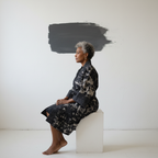 Woman sitting on a white block against a plain background with a black abstract shape.